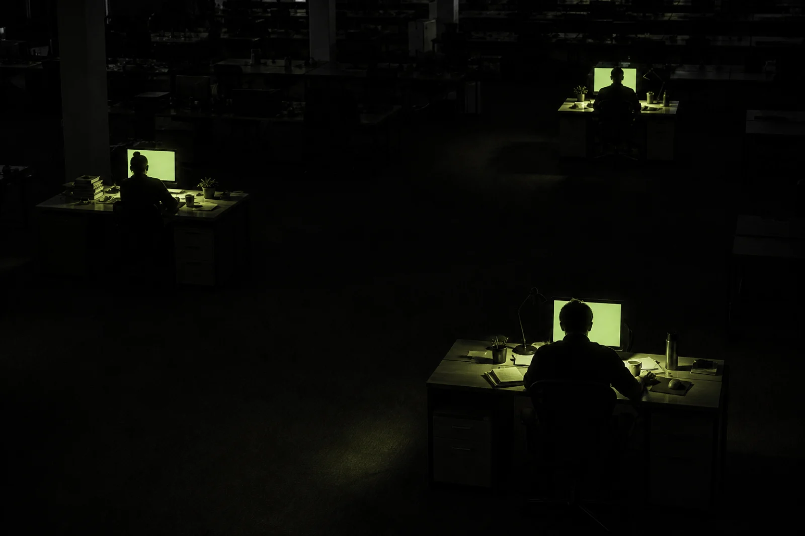 Three people at separate desks, each lit only by their own screen, the pools of light never touching